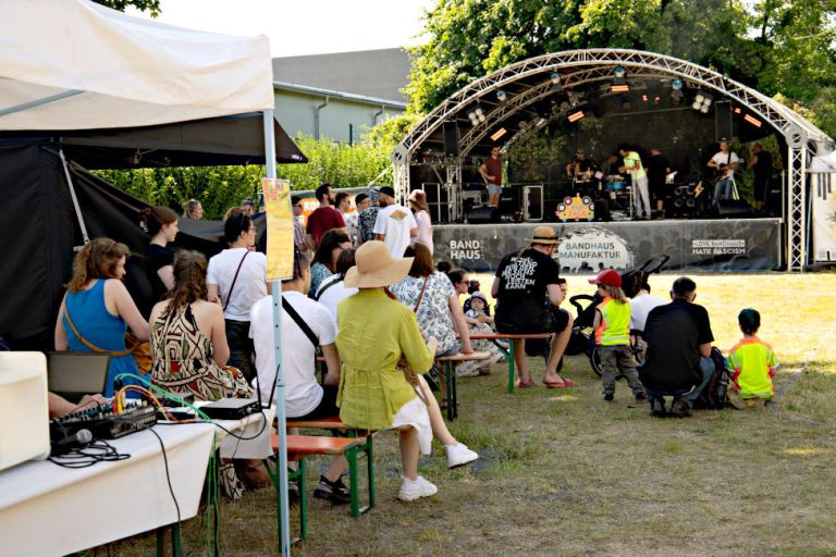 Auf eine wiese vor einer Bühne sitzen Menschen jeden Alters auf Bierbänken und sehen sich eine Konzert an.