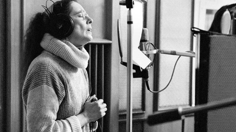 Schwarz-Weiß-Foto der Künstlerin, die einen Rollkragenpullover und eine Nickelbrille trägt, wie sie in einem Studio in das Mikrofon singt.