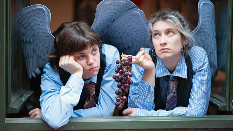 Zwei Schauspielerinnen sind als Engel verkleidet und schauen gelangweilt aus einem Fenster.