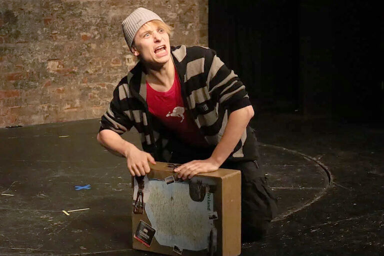 Ein Schauspieler kniet neben einem Koffer, schaut nach oben und schreit mit weit geöffnetem Mund.