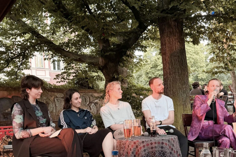 Fünf Teilnehmerinnen und Teilnehmer einer Diskussionsrunde sitzen hinter einem kleinen Tisch mit Getränken im Freien.