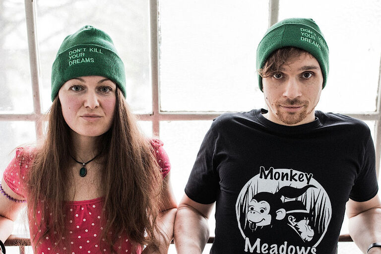 Foto des Duos in sommerlicher Kleidung, aber mit grünen Mützen vor einem altem Fabrikfenster