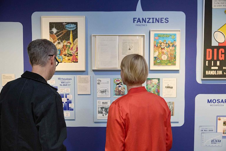 Ein Mann und eine Frau stehen vor einer Wandtafel und schauen sich Bilder und Informationen zum Mosaik an.