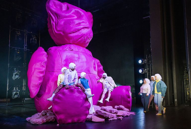 Mehrere Schauspielerinnen und Schauspieler sitzen auf oder stehen an einem berdimensionalen, pinkfarbenen Teddybär.