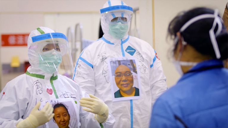 Eine Ärztin und ein Arzt stehen vollkommen verhüllt durch Schutzanzüge und Masken vor einer Patientin und haben Fotos von ihren Gesichtern auf der Brust, damit diese weiß, wie sie aussehen.