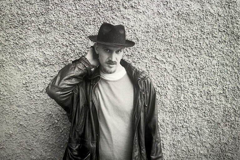 Schwarz-Weiß-Foto des Musikers mit Hut und Lederjacke vor einer grob verputzten Mauer