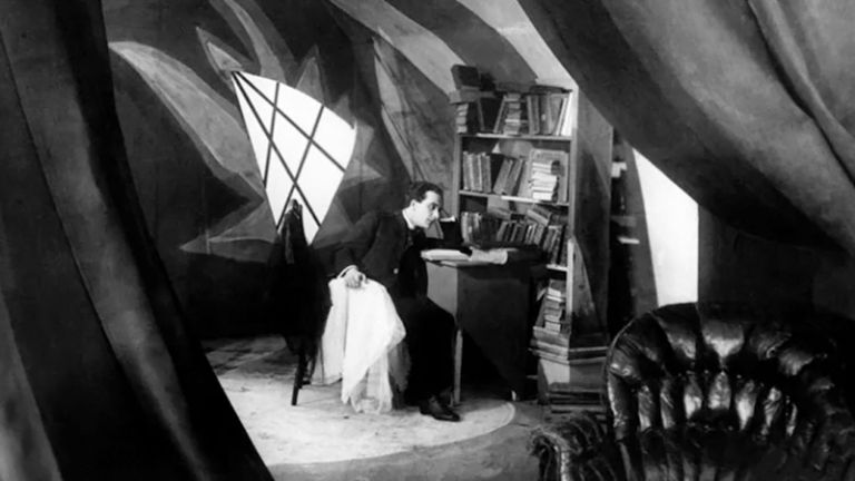 Filmszene, in der ein junger Mann im Anzug in einem futuristischen Arbeitszimmer am Schreibtisch sitzt und grübelt.