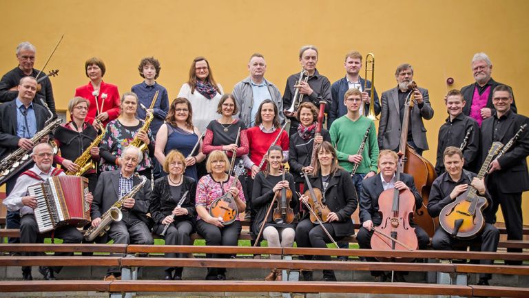 Gruppenfoto der Musiker*innen des Orchesters mit ihren Instrumenten auf den Bänken der Parkbühne GeyserHaus.