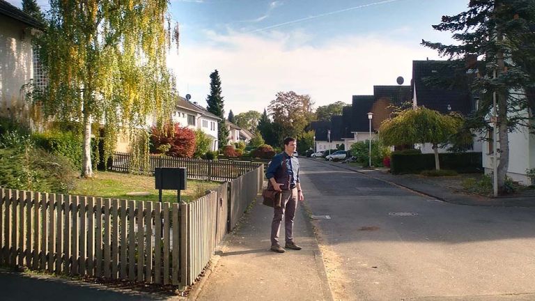 Ein junger Mann steht an einem sonnigen Tag allein auf der Straße einer gepflegten Siedlung mit Eigenheimen und Doppelhaushälften.