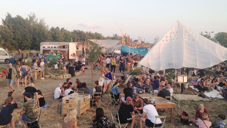 Viele Menschen sitzen an Holztischen oder auf Decken am Boden und genießen einen schönen Abend.