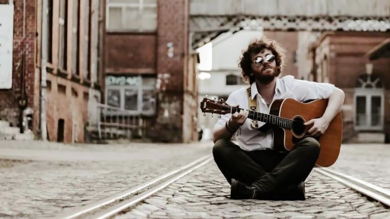 Der Musiker sitzt mit Sonnenbrille auf dem Boden und spielt Gitarre.