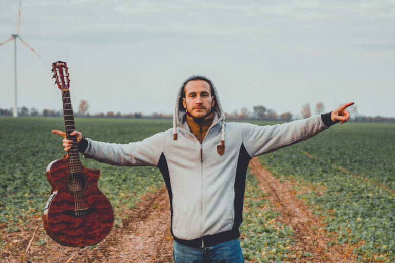 Der Künstler läuft an einem grauen Tag über ein abgeerntetes Feld, hebt beide Hände, wovon eine die Gitarre hält
