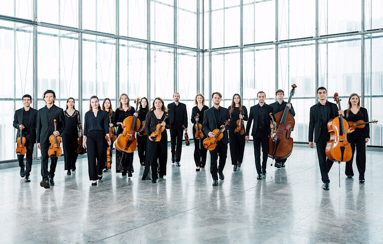 Gruppenbild des Orchesters mit seinen Instrumenten in einem großen Raum mit Glaswänden