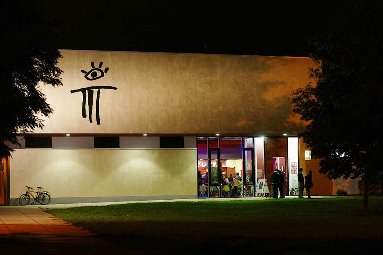 Außenansicht des Theaters bei Nacht, in dessen hell erlauchteten Foyer viele Menschen sind