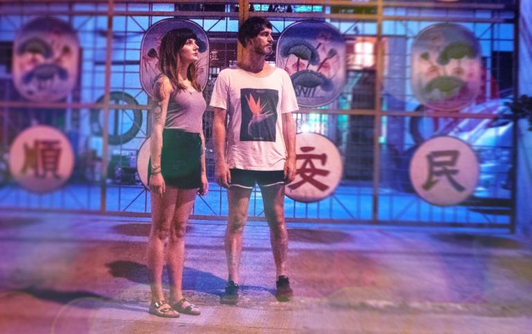 Das Duo steht vor einem Gitter, an dem asiatische Schreiftzeichen angebracht sind.