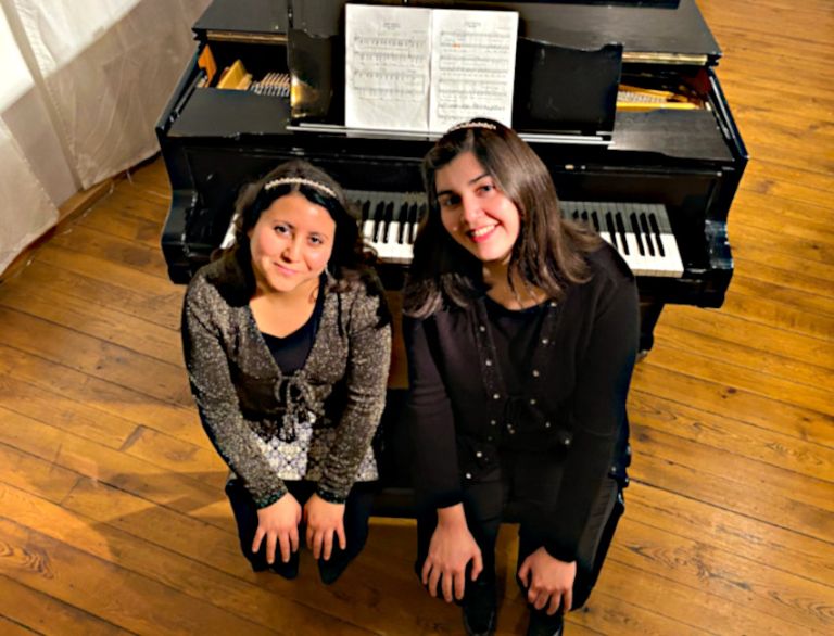 Die beiden Pianistinnen sitzen freundlich lächelnd nebeneinander auf einer Bank mit dem Rcken zu ihrem Konzertflgel.