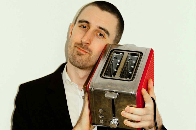 Der Musiker hält sich lächelnd einen Toaster an die Wange.
