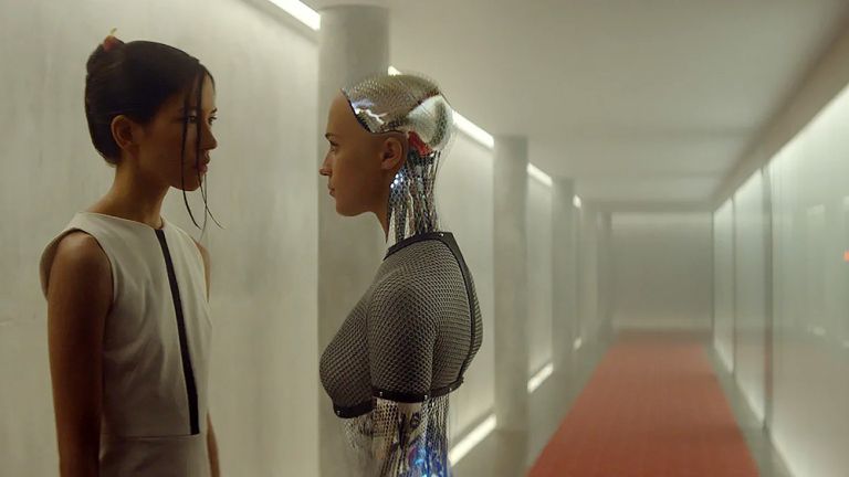 Eine Frau und ein weiblich scheinender Roboter stehen sich in einem kahlen Gang gegenüber.