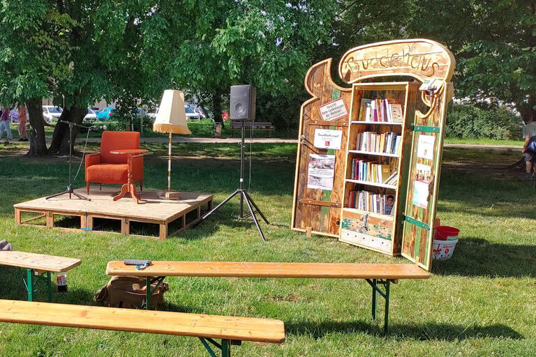 In einem kleinen Park stehen Bänke, ein Bücherschrank und ein Podium mit Sessel