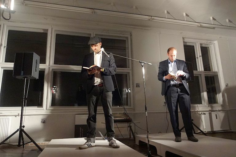 Zwei Männer in schwarzen Anzügen stehen nebenainander auf niedrigen Podesten, halten Bücher in den Händen und lesen daraus.