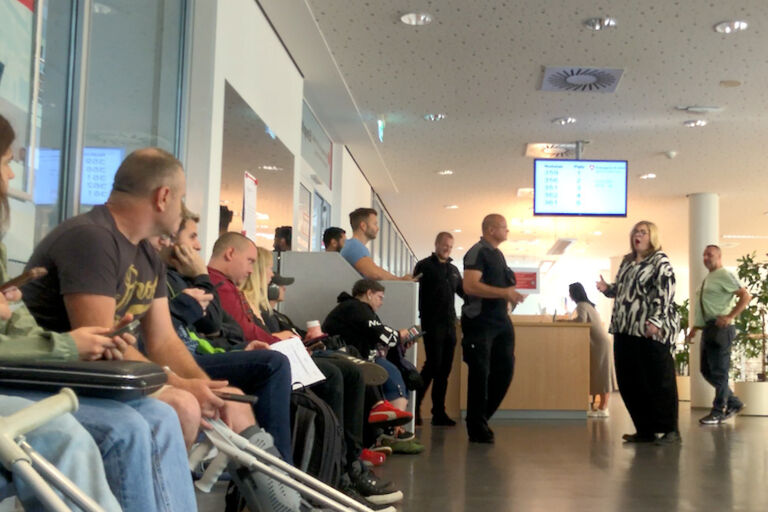 Eine Opernsängerin singt ein einem Wartebereich vor vielen Personen, die auf Bänken sitzen oder im Raum stehen.