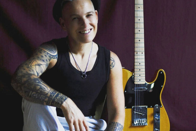 Die Musikerin sitzt lächelnd vor ihrer Gitarre, die an der Wand lehnt, auf dem Boden.