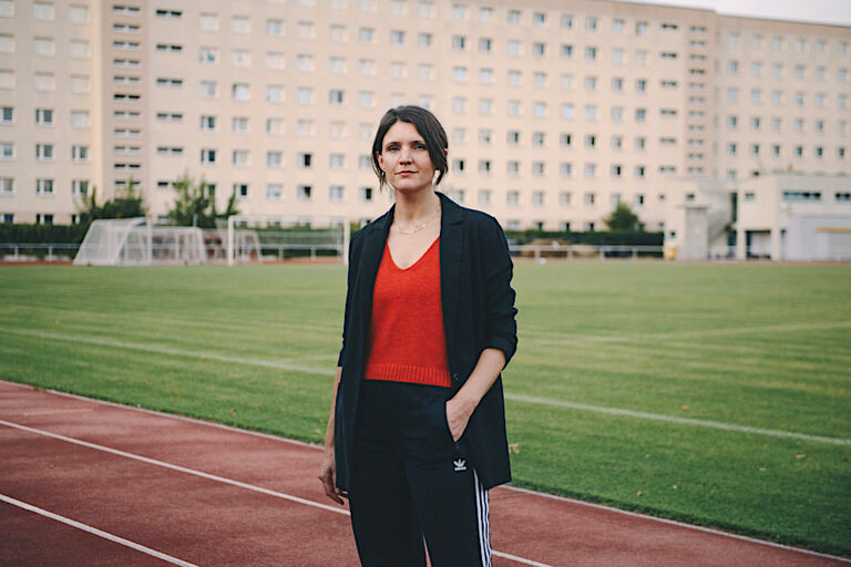 Die Schriftstellerin steht in einer Jogginghose auf einem Sportplatz vor einem Plattenbau.