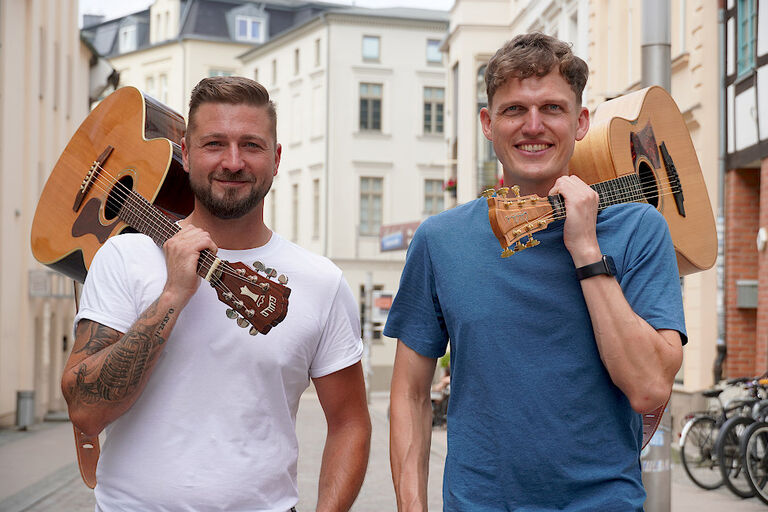 Die beiden Musiker stehen mit ihren Gitarren auf der Schulter lächelnd nebeneinander.