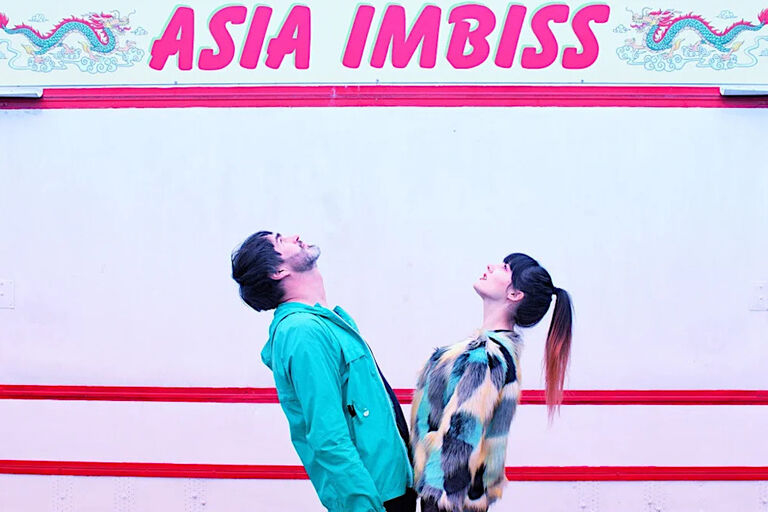 Ein Mann und eine Frau schauen nach oben zu einem Schild mit der Aufschrift "Asia Imbiss".