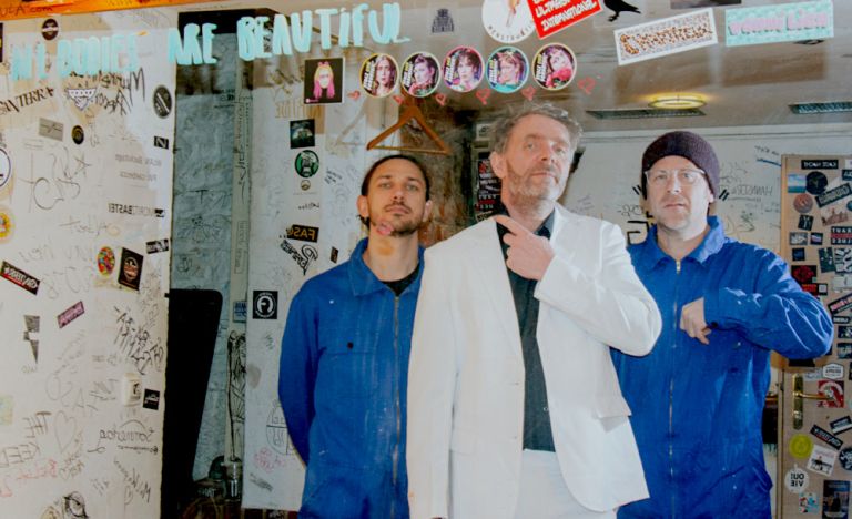 Gruppenfoto, auf dem der Sänger einen weißen Anzug trägt und die beiden anderen Musiker einen blauen Overall