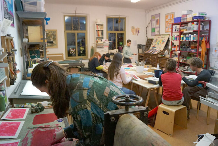 Viele Kinder sitzen an einem Tisch voller bunter Blätter. Im Vordergrund arbeitet eine Frau an einer Druckerpresse.