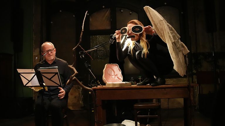 Ein Schauspieler sitzt neben einem Tisch und liest seinen Text vor, während neben ihm auf dam Tisch eine Schauspielerin hockt, die einen Raben spielt.