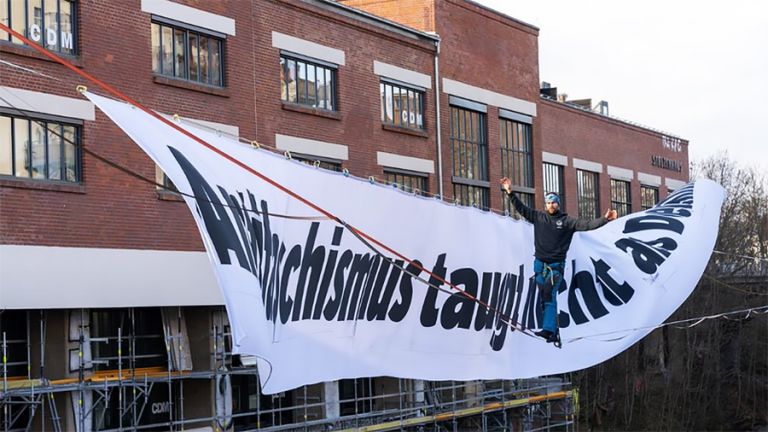 Ein großes Banner mit der Aufschrift "Antifaschismus taugt nicht als Denkmal", vor dem ein Mann auf einer Slackline balanciert