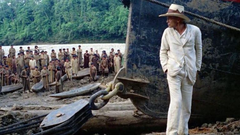 Klaus Kinski als Fitzgerald steht im weißen Anzug vor einem alten Dampfboot. Im Hintergrund sieht man einen Fluß, den Urwald und eine große Gruppe von indigenen Menschen.