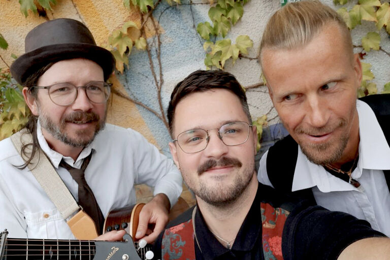 Selfie der drei Musiker vor einer bunten Wand mit Efeu-Ranken