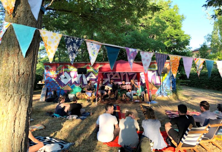 Viele Menschen sitzen auf Decken oder Liegestühlen vor einer kleinen Bühne im Park.