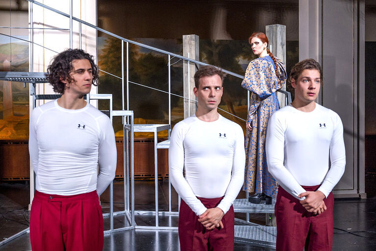 drei junge Männer stehen in Sportkleidung in einer Reihe. Im Hintergrund ist eine Frau im langen Kleid zu sehen.