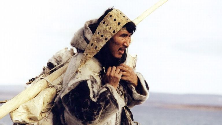 Foto von Atanarjuat, der in traditioneller Kleidung in einer weiten Landschaft unterwegs ist und ein langes Stck Elfenbain auf den Schultern trägt.