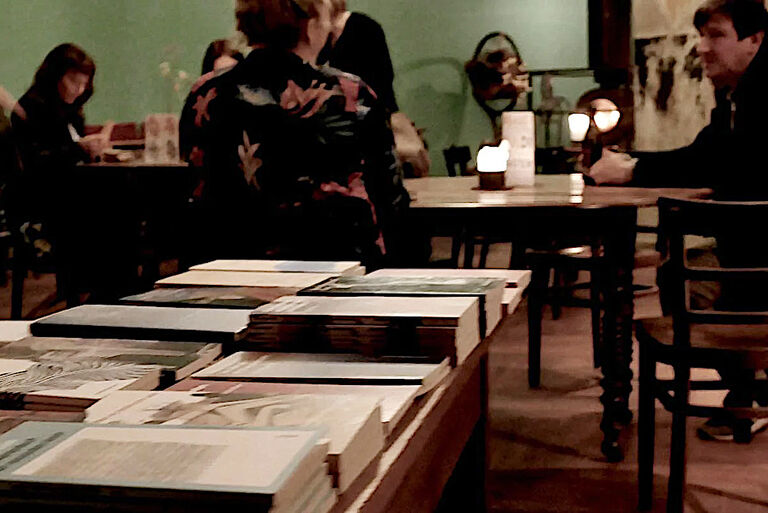 Hinter einem Büchertisch sitzen an weiteren Tischen mehrere Personen in einer Bar.