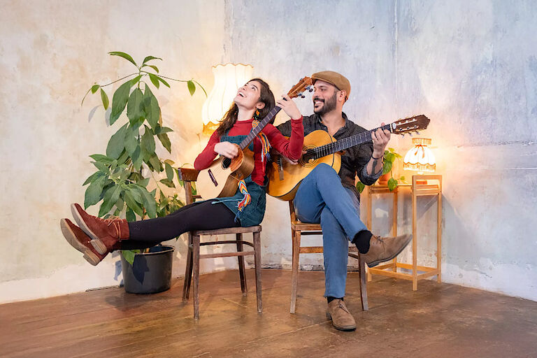 Das Duo sitzt mit seinen Instrumenten lachend auf Sthlen in einer Zimmerecke.