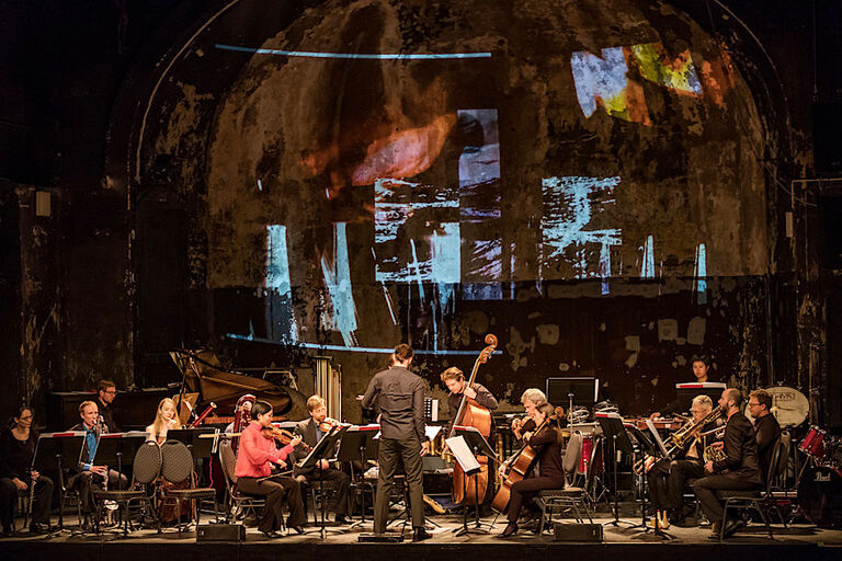 Das Ensemble bei einem Konzert auf einer großen Bühne auf deren hintergrund ein abstraktes Bild projeziert wird