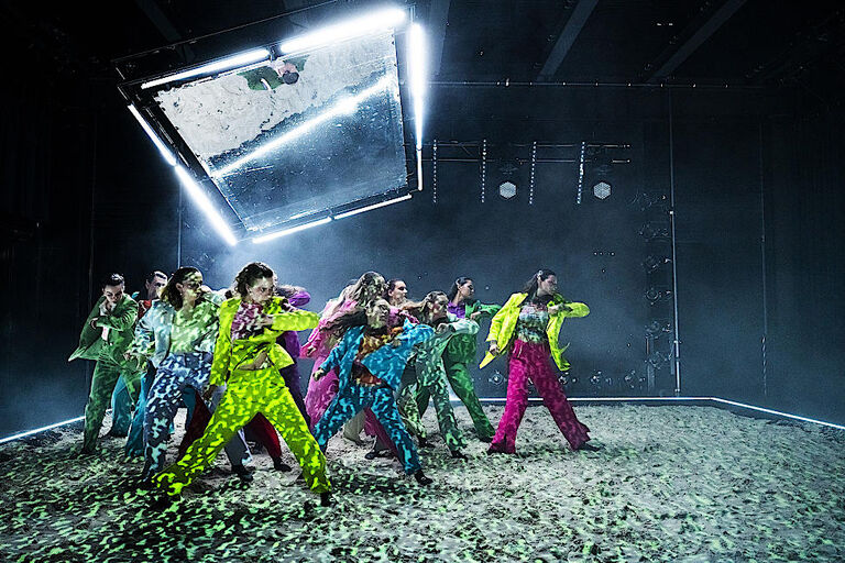 Eine Gruppe junger Tänzerinnen und Tänzer in neonfarbigen Kostümen tanzen in einer Formation.