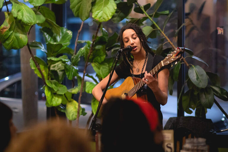 Die Künstlerin sitzt bei einem Auftritt mit Gitarre vor einer großen Grünpflanze.