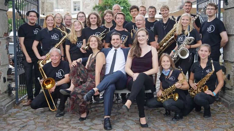 Das große fröhliche Ensemble der Bigband mit Instrumenten auf einem Gruppenfoto