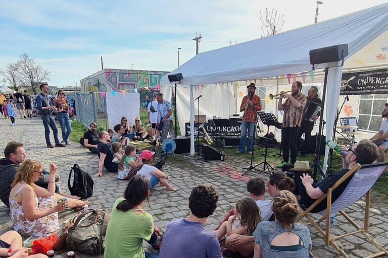 Kleine und große Zuschauer sitzen in Liegestühlen oder auf dem boden und lauschen einem Konzert beim Stadtteilfest.