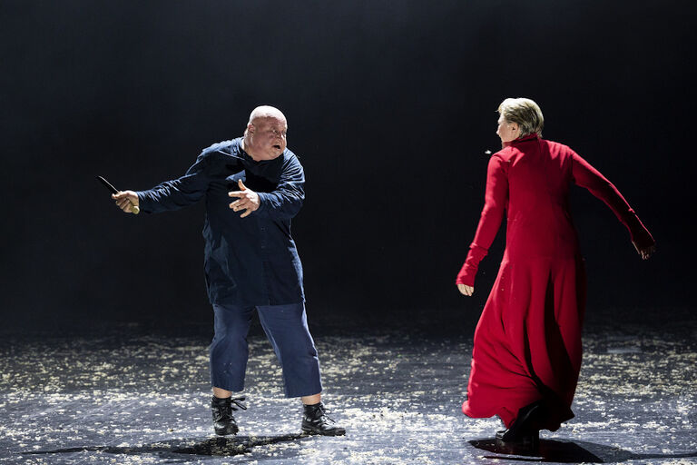 Ein schauspieler mit einem Messer und eine Frau in einem roten Mantel stehen sich auf der Bühne gegenüber.
