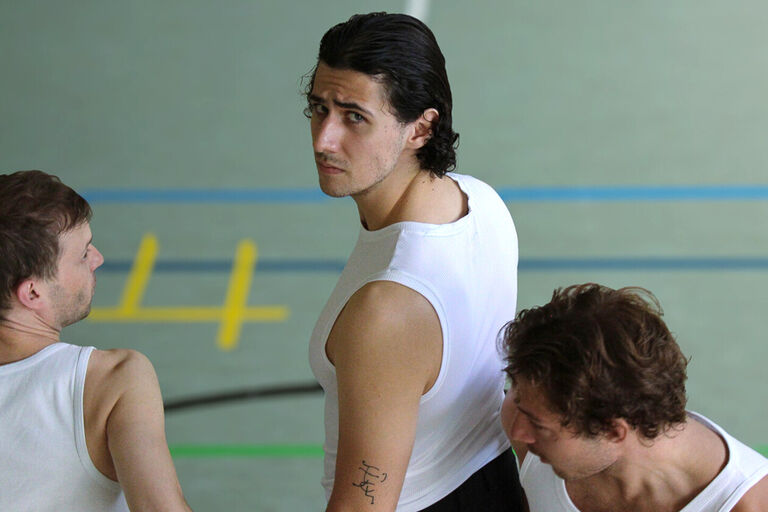 Drei junge Männer sind in einer Sporthalle. Der mittlere von ihnen schauf mit traurigem Blick zurück.