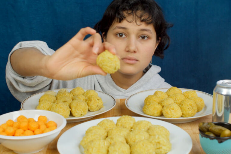 Eine junge Frau an einem Tisch mit gefüllten Tellern und Schüsseln hält etwas hoch, das wie eine Teigbällchen aussieht.