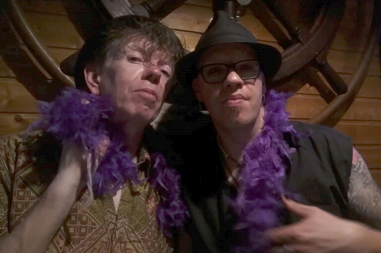Foto der beiden Künstler mit einer Federboa in Lila um den Hals.