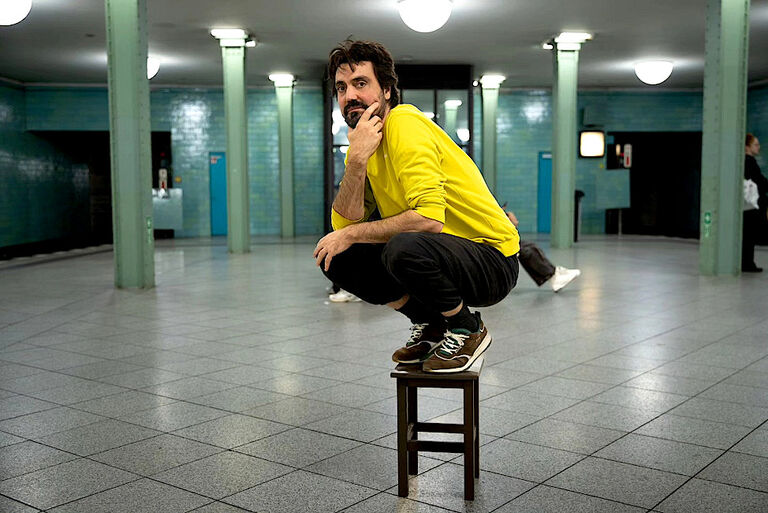 Der Musiker hockt in einer U-Bahnstation auf einem kleinen Holzhocker.